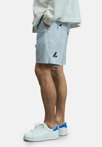 Shorts de sport gris clair avec poches latérales, ornés d'un logo noir, associés à des baskets blanches accentuées de bleu. Texture de tissu lisse.