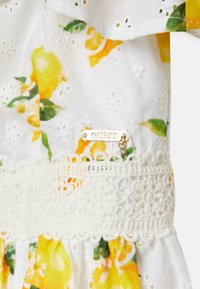 Weißes Baumwollkleid mit gelben Blumenmustern, verziert mit einer Spitzenakzent in der Taille und einem goldenen "GUESS"-Tag, der mit einem Charme befestigt ist.