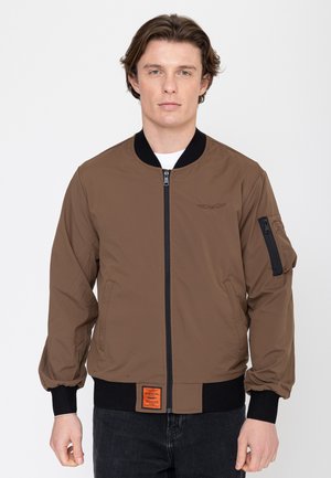 Junger Mann trägt eine braune Bomberjacke mit schwarzem Kragen, Bündchen und Reißverschluss, steht vor einem schlichten weißen Hintergrund.
