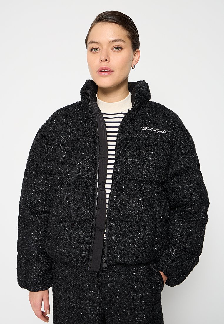 Schwarze Pufferjacke mit strukturiertes Oberfläche, Reißverschluss, hohem Kragen und besticktem Logo. Gestreiftes Hemd darunter getragen.