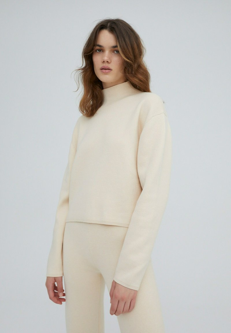 Crème-kleurige cropped sweater met een hoge hals, gemaakt van een zachte stof. Heeft lange mouwen en ribgebreide randen voor textuur.