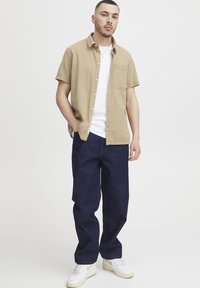 Korte mouw khaki overhemd met textuur, wit T-shirt met ronde hals, donkerblauwe broek met een ruime pasvorm en witte sneakers met gele accenten.