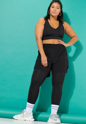 Una mujer luce ropa deportiva negra, calcetines a rayas y zapatillas blancas sobre un fondo de estudio turquesa.