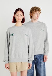 Grijze, lange mouwen sweatshirts met groene grafische accenten, geribbelde manchetten en halslijn. De ene model draagt shorts, de andere jeans.