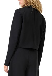 Sieviete ar blondiem matiem, tērpusies pieguļošā melnā jakā ar īsiem piedurknēm un atbilstošās augstās jostasvietas biksēs, redzama no aizmugures uz balta fona.