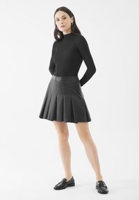 Gonna plissettata nera in pelle con vita aderente, abbinata a un top nero a maniche lunghe e mocassini neri, su sfondo bianco.