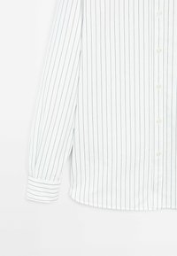 Chemise blanche à manches longues boutonnée avec fines rayures verticales noires et poignets rayés assortis sur les manches.