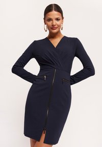 Granatowa sukienka z długim rękawem, z dekoltem w kształcie litery V, z przednimi kieszeniami zapinanymi na zamek oraz rozcięciem sięgającym ud na dole. Wykonana z elastycznego materiału.