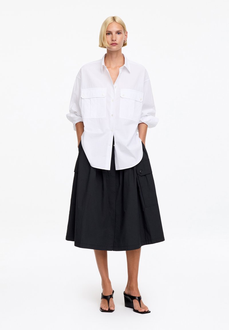 Femme aux cheveux blonds courts porte une chemise blanche boutonnée aux manches retroussées, une jupe midi noire à poches et des sandales à talons noires.