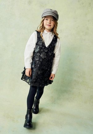 Schwarzes Blumenkleid über einem weißen Kragenhemd, kombiniert mit schwarzen Strumpfhosen und klobigen schwarzen Stiefeln. Eine graue Mütze ergänzt das Outfit.