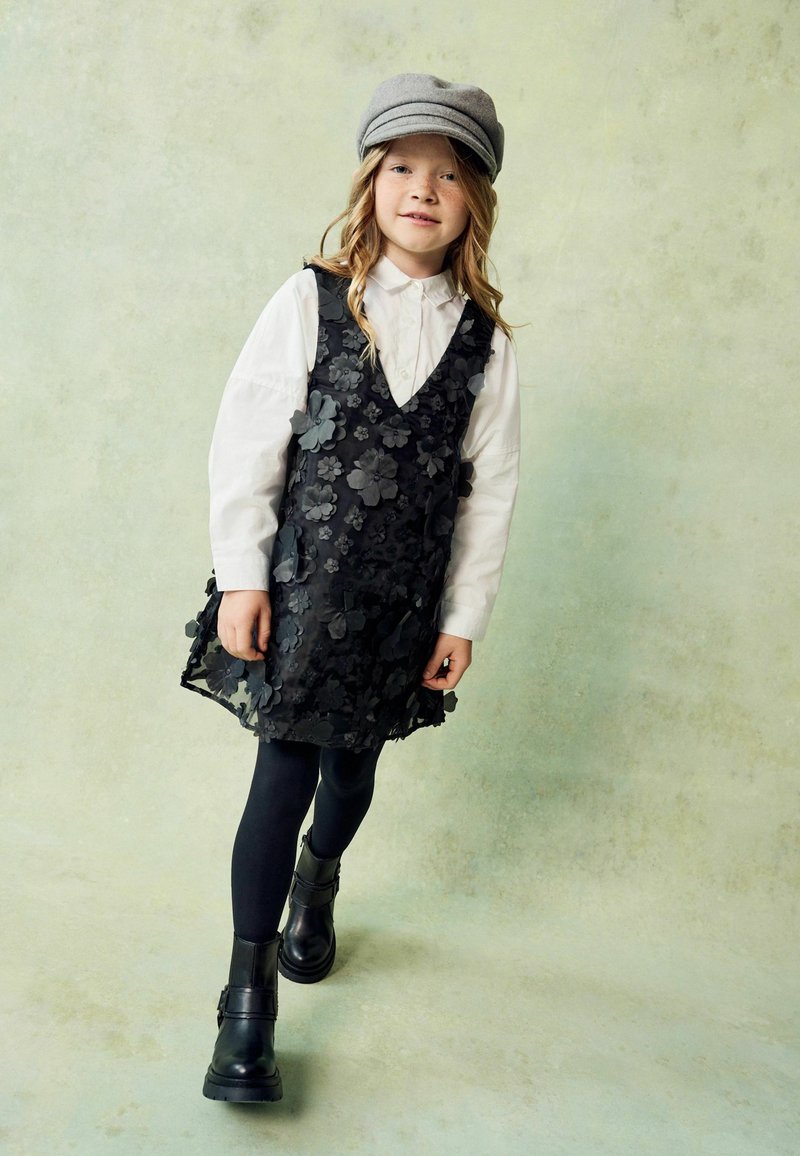 Schwarzes Blumenkleid über einem weißen Kragenhemd, kombiniert mit schwarzen Strumpfhosen und klobigen schwarzen Stiefeln. Eine graue Mütze ergänzt das Outfit.
