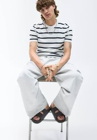 Model iført en hvid t-shirt med marineblå striber, grå oversized sweatpants og mørke slidesandaler, siddende på en metalstol.