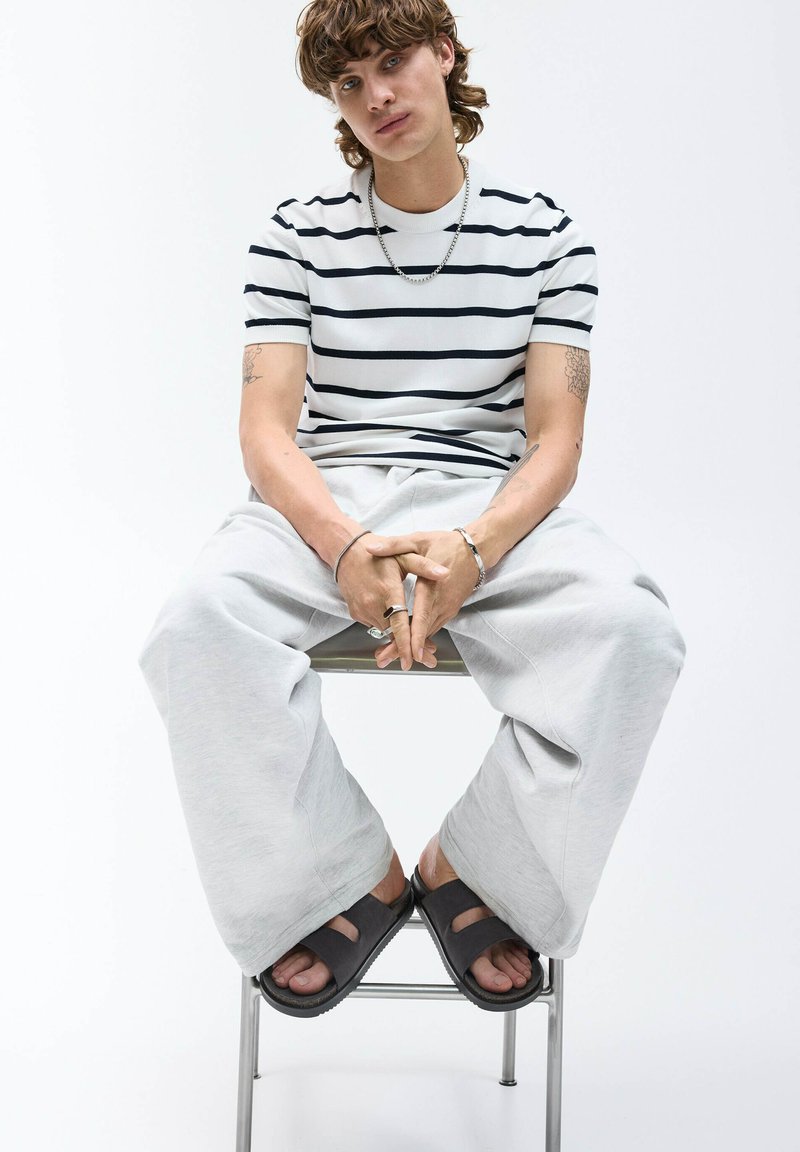 Model iført en hvid t-shirt med marineblå striber, grå oversized sweatpants og mørke slidesandaler, siddende på en metalstol.