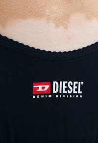 Svart tyg linne med ett scallop-mönster i halsen och en röd och vit "DIESEL DENIM DIVISION" logotyp tryckt på framsidan.