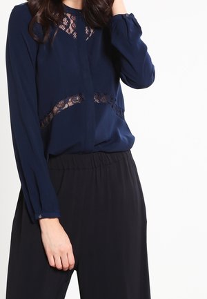 Femme portant un chemisier bleu marine avec des détails en dentelle sur la poitrine et la taille, associé à un pantalon noir ample, sur un fond blanc uni.