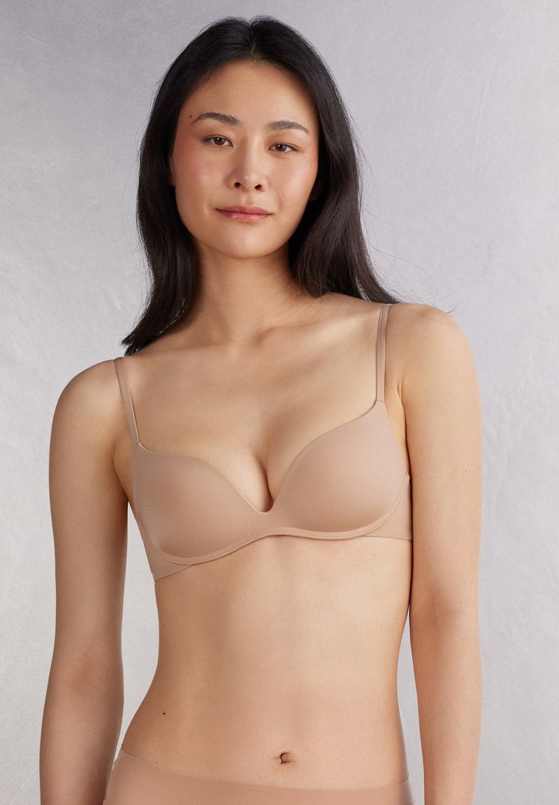 Sostén con escote en pico en tono nude, con copas suaves, tirantes ajustables y bordes sin costuras. La tela parece suave y ajustada contra la piel.