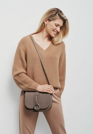 Beżowy sweter dziany z dekoltem w kształcie V, luźnym kroju oraz prążkowanymi mankietami, w połączeniu z brązową torebką na ramię z okrągłym metalowym akcentem.