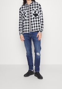 Czarno-biała flanelowa koszula w kratę z dwoma kieszeniami na piersi, noszona na szarej koszulce, zestawiona z podartymi niebieskimi dżinsami i czarnymi trampekami.