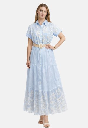 Donna che indossa un abito lungo ricamato azzurro chiaro con maniche corte, cintura intrecciata e tacchi aperti beige, posa con la mano sul fianco.
