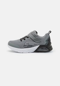 Chaussure de sport grise avec une tige en mesh texturé, des accents noirs, une sangle ajustable et une semelle blanche avec un coussin d'air visible.