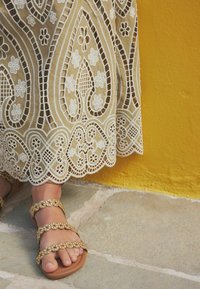Robe crème ornée de dentelle avec des motifs floraux et paisley complexes, associée à des sandales brunes dotées de lanières avec des accents dorés.