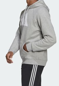 Sudadera gris claro con capucha, bolsillo tipo canguro y logotipo blanco en el pecho. Cuenta con puños elásticos y una tela suave y texturada.
