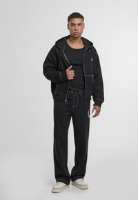 Sudadera negra con costuras blancas en contraste y cremallera frontal, combinada con pantalones de chándal negros con ajuste de cordón y logo lateral.