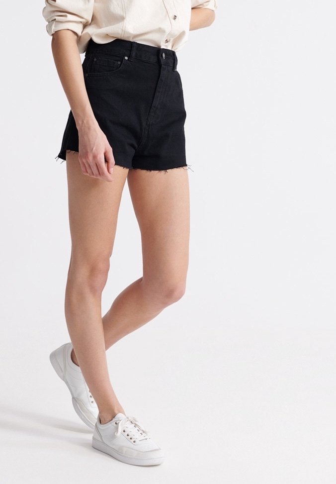 Black denim shorts with frayed hem, high-waisted design. Paired with white sneakers, showcasing a minimalist style in a neutral background.