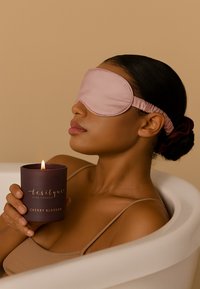 Masque de sommeil en soie rose, peau brune, tenant une bougie violette foncée étiquetée "Fleur de cerisier" avec une mèche allumée, assise dans une baignoire blanche.