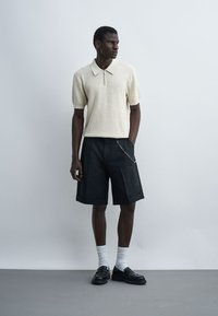 Homme portant un polo texturé à manches courtes, un short noir au genou avec une chaîne, des chaussettes blanches et des mocassins noirs, debout devant un fond uni.