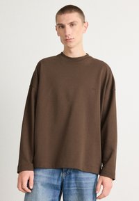 T-shirt marron à manches longues avec une coupe décontractée, col rond et discret logo. Associé à un jean en denim bleu clair.