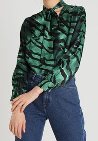 Green and black patterned blouse with a bow tie neckline, long sleeves, and a smooth texture, paired with high-waisted blue denim jeans.