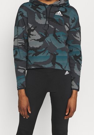 Person wearing a blue, gray, and black camouflage Adidas hoodie and black Adidas leggings, standing with arms relaxed by their sides.