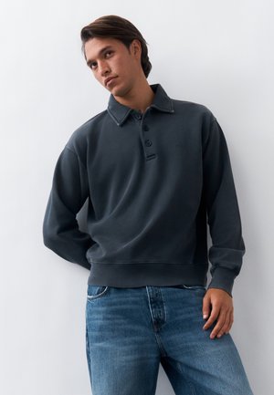 Νέος άνδρας που φοράει σκούρο γκρι πουλόβερ με κουμπιά και μπλε τζιν, στέκεται μπροστά σε λευκό απλό φόντο.