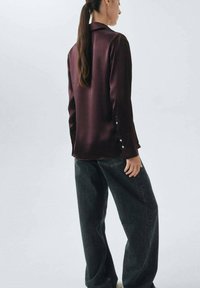 Camisa de satén burdeos con cuello, mangas largas y detalles de botones; combinada con jeans de mezclilla oscuros. Textura suave y corte relajado.