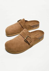 Barna velúr klumpák csattal, parafa részletezéssel a sarkon és texturált gumitalppal. Kerek orrú és nyitott hátú design.