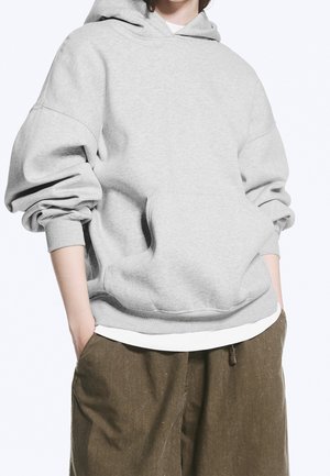 Personne portant un sweat à capuche oversize gris clair avec une poche kangourou et un pantalon marron ample, les mains dans les poches.
