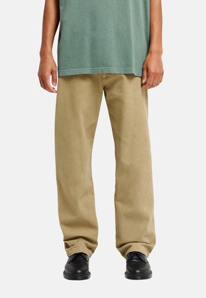 Person trägt lockere beige Hose, schwarze Schuhe und ein grünes Hemd mit entspannten Händen an den Seiten vor einem einfarbigen hellen Hintergrund.