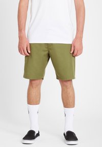 Mand iført olivengrønne shorts, hvide crew-sokker, sorte slip-on sko og en hvid t-shirt, der står foran en hvid baggrund.