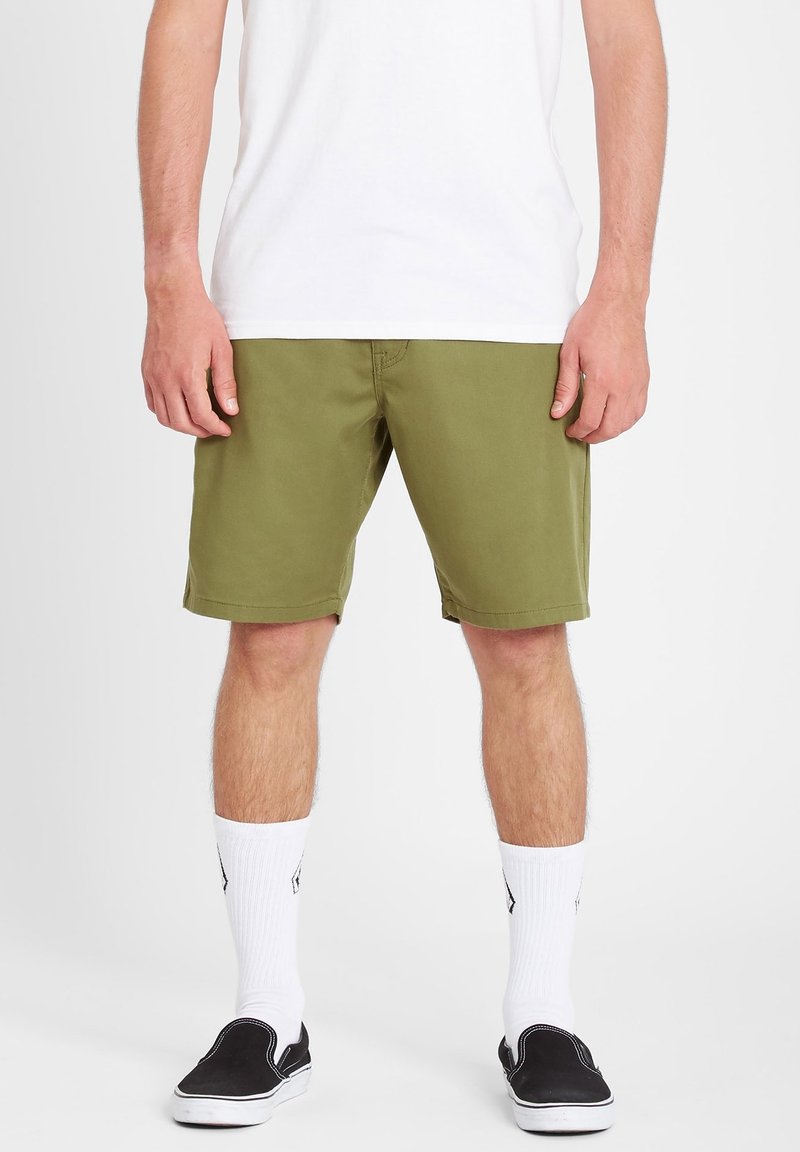 Mand iført olivengrønne shorts, hvide crew-sokker, sorte slip-on sko og en hvid t-shirt, der står foran en hvid baggrund.