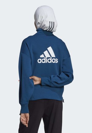 Person iført en blå Adidas-sweatshirt med logo på ryggen og en hvid hovedbeklædning med Adidas-striber, stående med den ene arm bag ryggen.