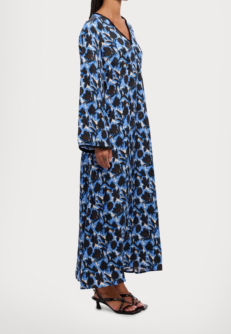Femme portant une longue robe fleurie bleue et noire à manches longues et des sandales à talons noires à lanières, debout de profil.