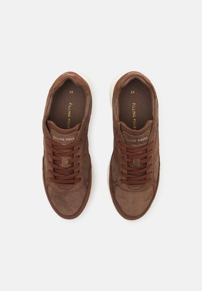 Pár hnědých semišových tenisek s tlustými bílými podrážkami a hnědými tkaničkami, fotografovaných shora na bílém pozadí.