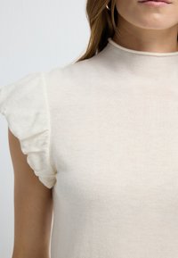 Haut crème avec des manches courtes à volants, col haut et un matériau doux et texturé. Design simple et ajusté.