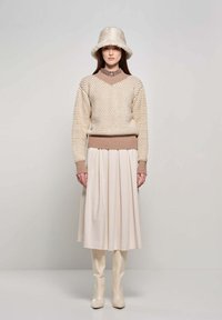 Maglione lavorato a maglia beige con motivo testurizzato, scollatura alta e polsini a coste, abbinato a una gonna plissettata crema e stivali lucidi al ginocchio.