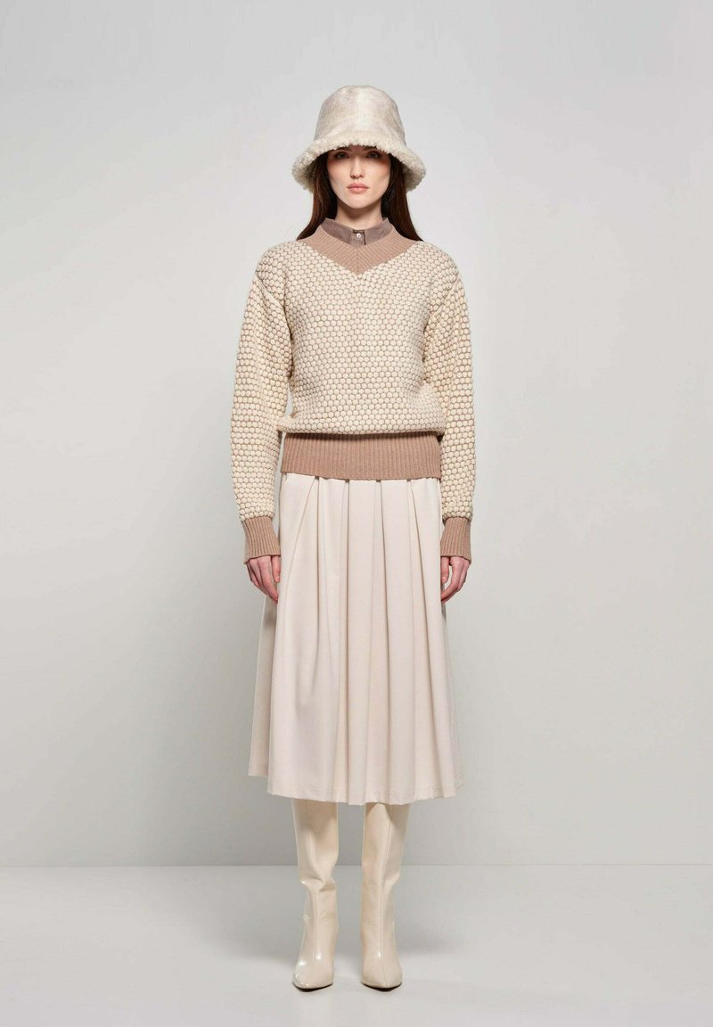 Maglione lavorato a maglia beige con motivo testurizzato, scollatura alta e polsini a coste, abbinato a una gonna plissettata crema e stivali lucidi al ginocchio.