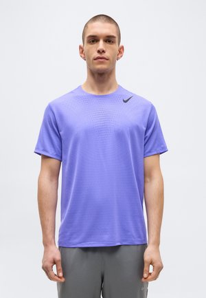 Man with short hair wearing a purple Nike dotted athletic shirt and gray shorts, standing against a plain white background.