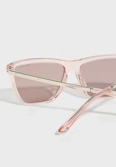 Διαφανή ροζ γυαλιά ηλίου σχήματος γατίσιου ματιού με σκουρόχρωμους φακούς και μεταλλικά μπράτσα, εκτεθειμένα σε λευκή επιφάνεια.