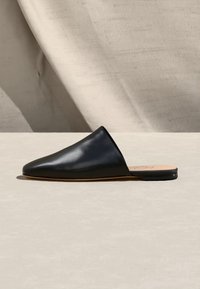 Μαύρο δερμάτινο mule χωρίς κορδόνια με μυτερή μύτη και επίπεδη σόλα, τοποθετημένο σε ουδέτερη επιφάνεια με φόντο μπεζ ύφασμα.