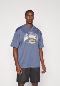 Grå nät basketttröja med "LOS ANGELES LAKERS" i gula och lila bokstäver. Har korta ärmar och en v-ringning.